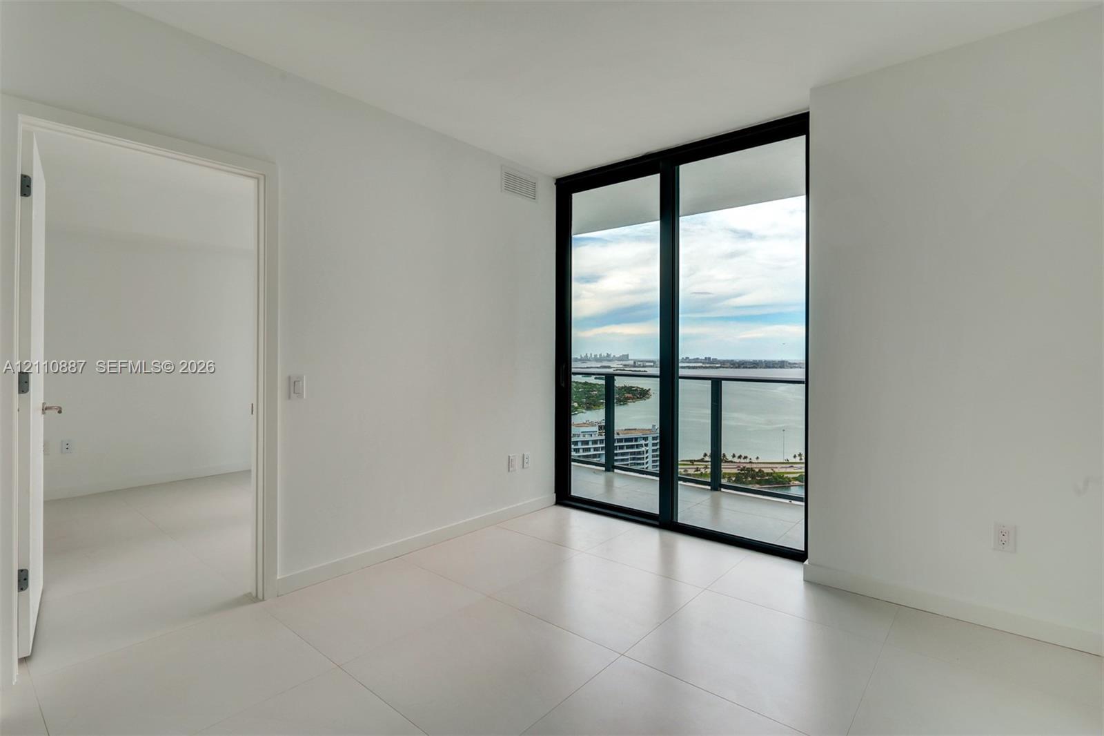 Floor-to-ceiling sliding glass doors, private balcony with metal railing, light-colored tile flooring, and neutral white wall finishes