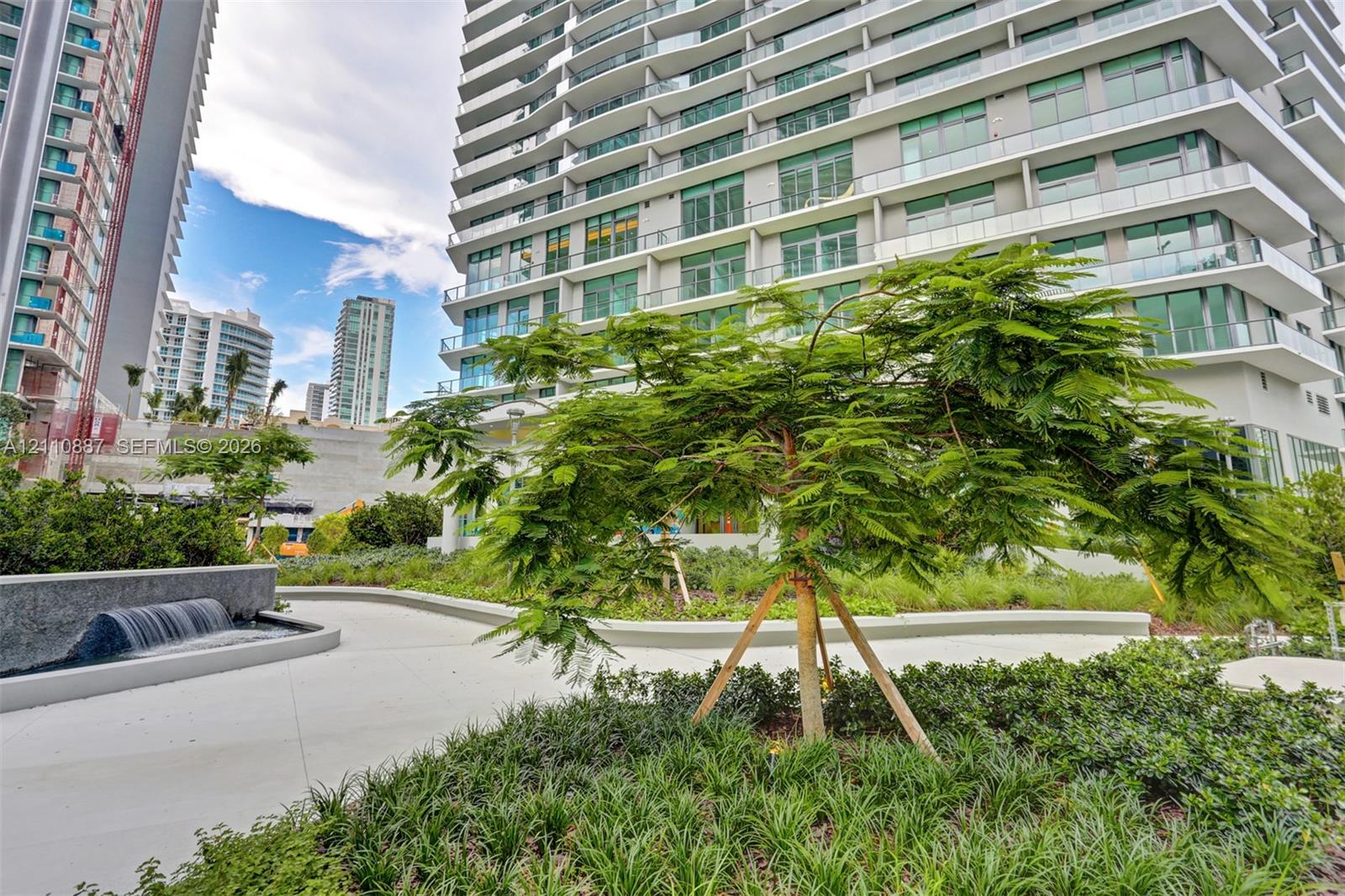 Landscaped courtyard featuring a tiered water feature, concrete walkways, mature plantings, and ornamental trees