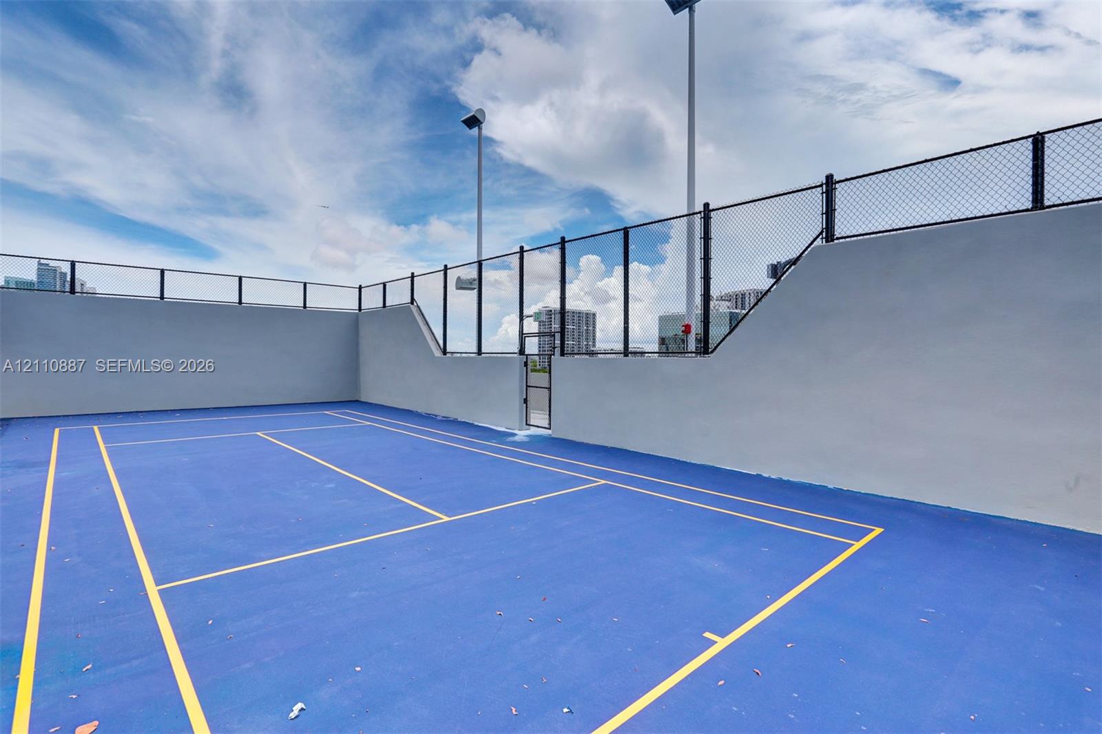 Recreational court featuring a high-contrast surface, perimeter chain-link fencing, solid masonry walls, and integrated court lighting