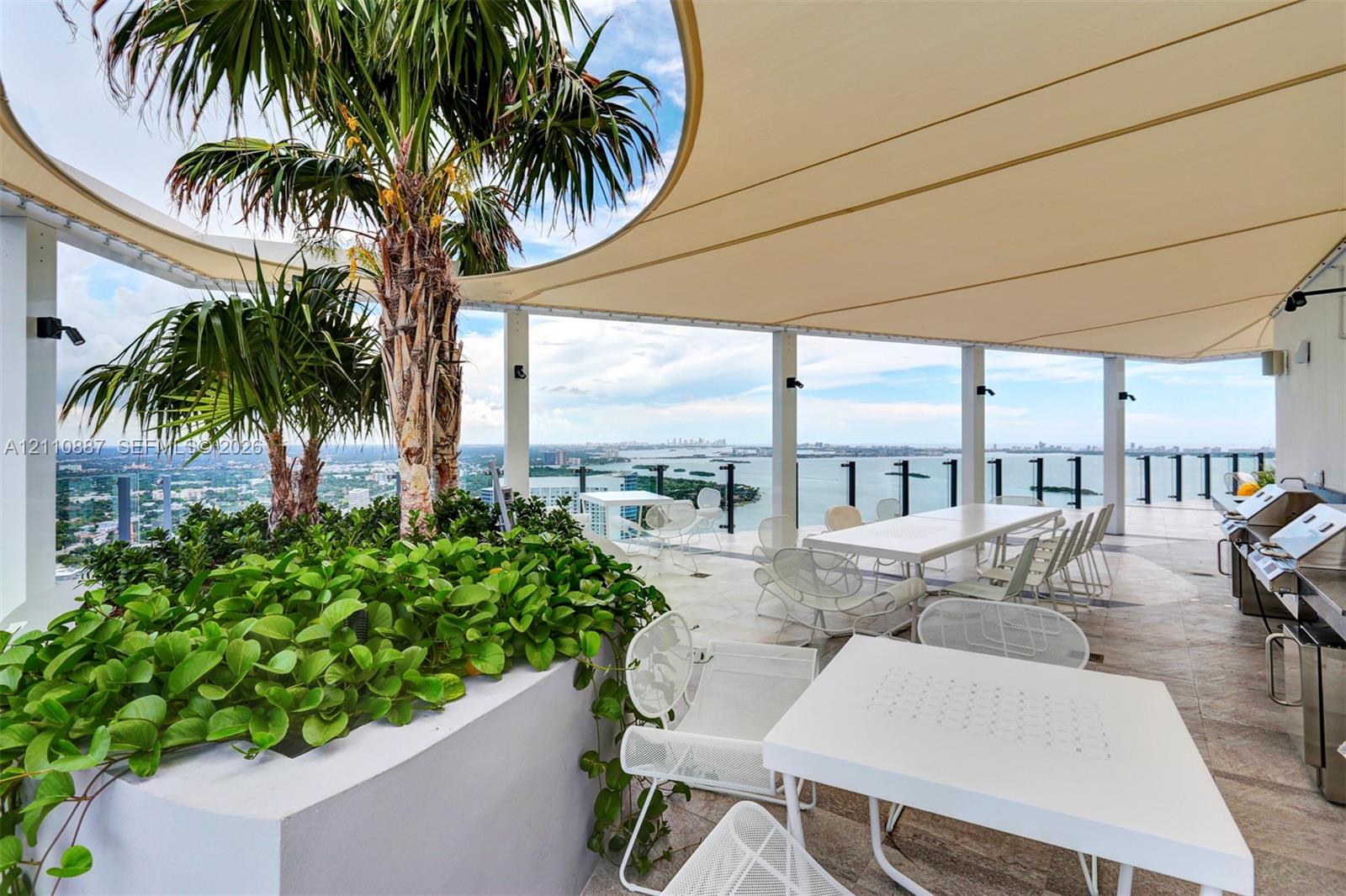 Covered rooftop terrace featuring large-format stone tile flooring, a custom-fitted tension shade structure, built-in concrete planter beds, and a stainless steel outdoor kitchen station