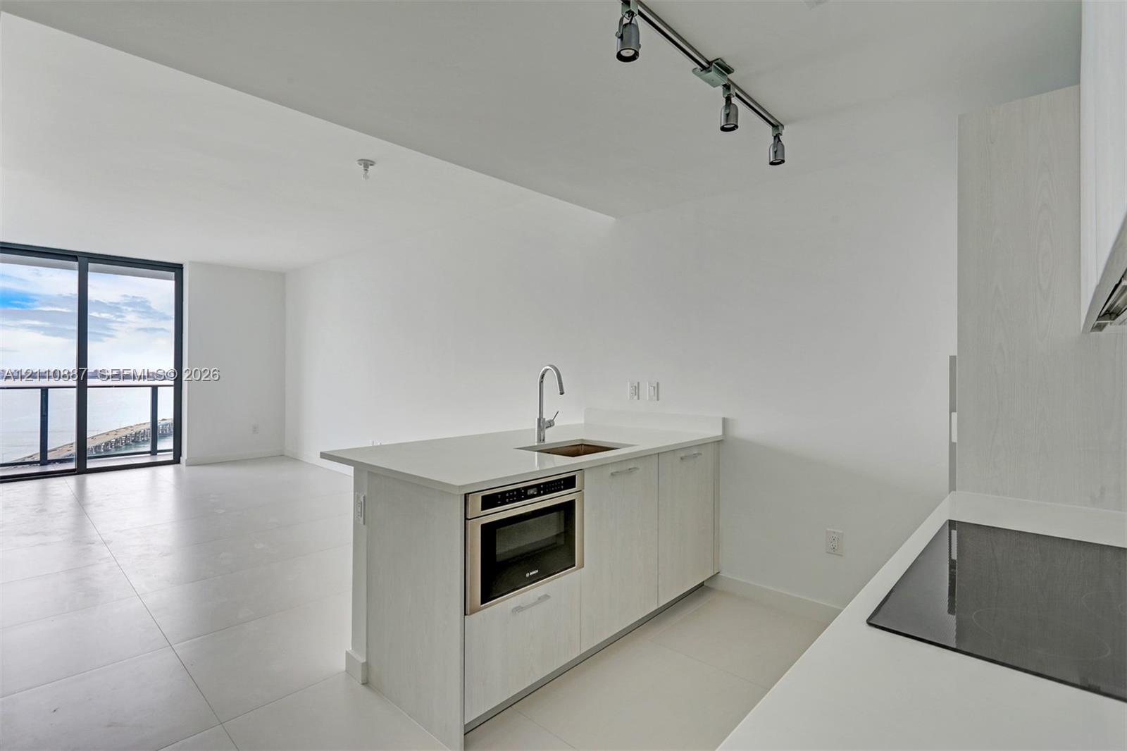 Kitchen featuring light-toned cabinetry, solid-surface countertops, built-in microwave, flat-panel cooktop, and track lighting
