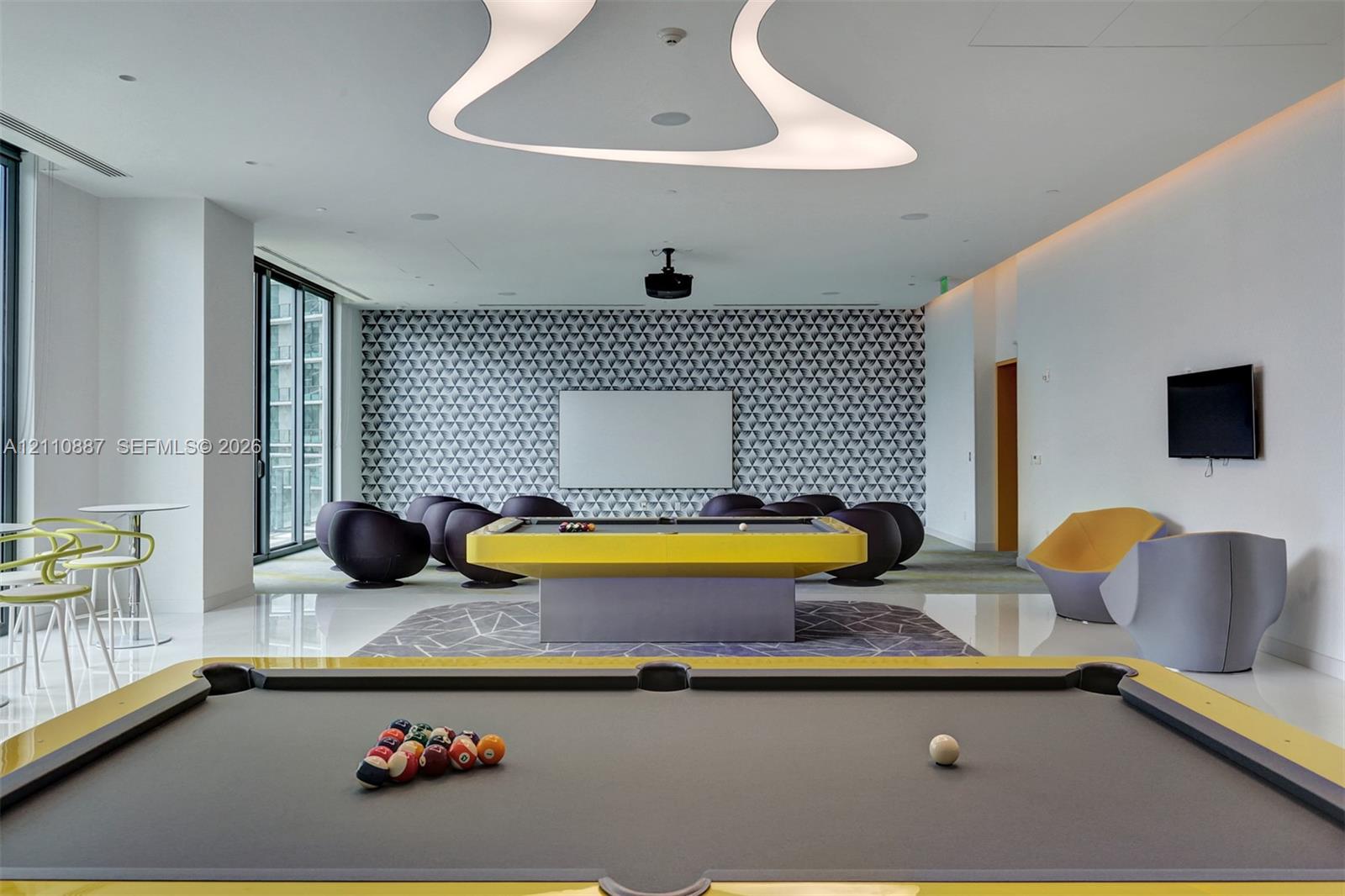 Recreation room featuring an abstract recessed ceiling light fixture, patterned accent wall, sleek flooring, and floor-to-ceiling glass paneling