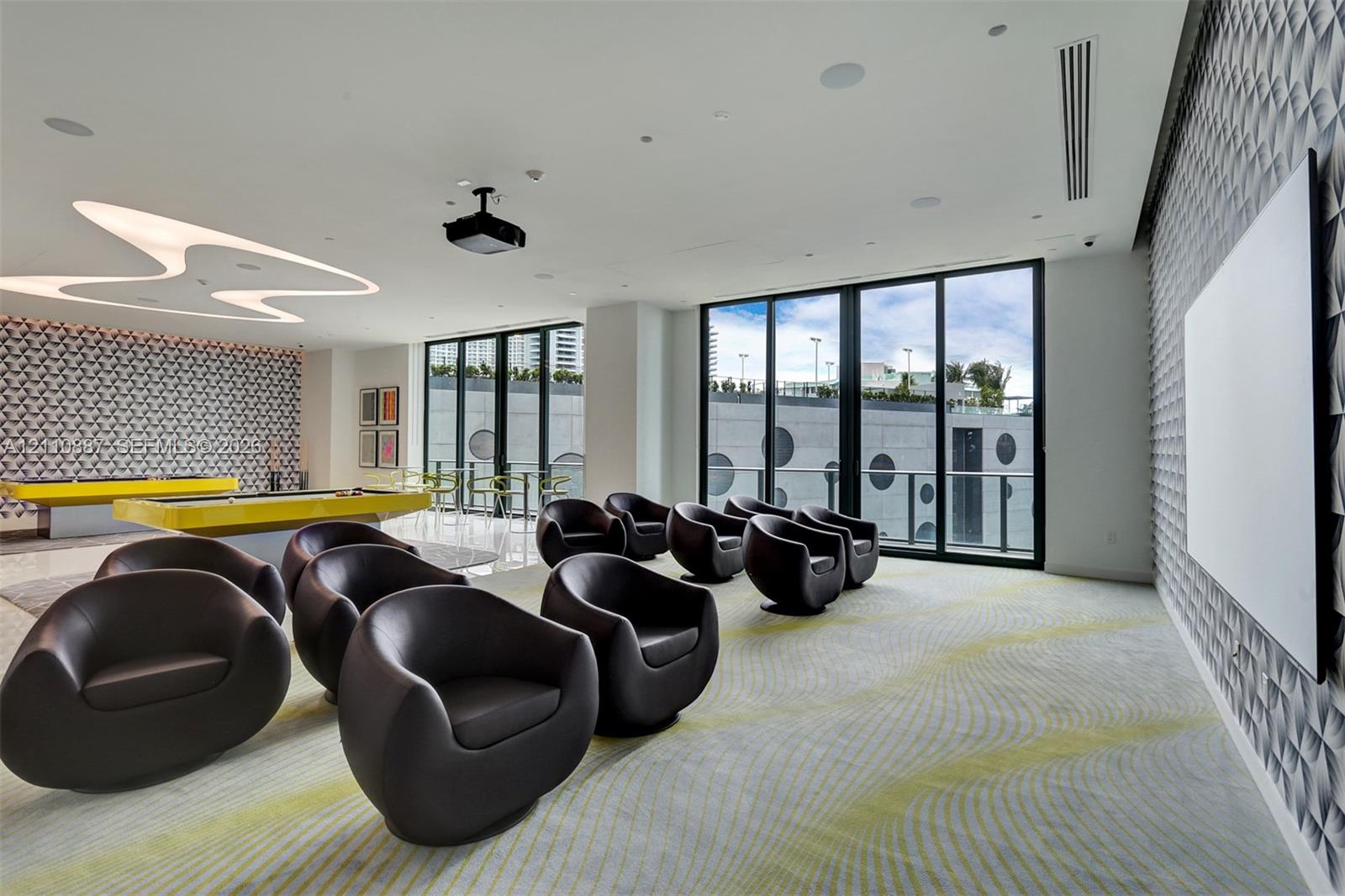 Multi-purpose room featuring textured wall panels, floor-to-ceiling glass sliding doors, custom overhead sculptural lighting, integrated ceiling speakers, and a ceiling-mounted projector