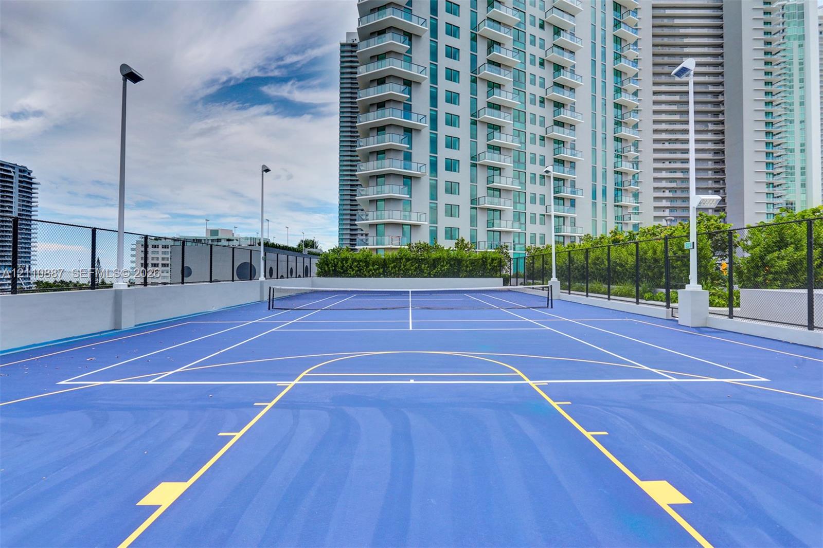Multi-use sports court with a blue surface, painted court markings, permanent net system, high-pole lighting, and perimeter fencing