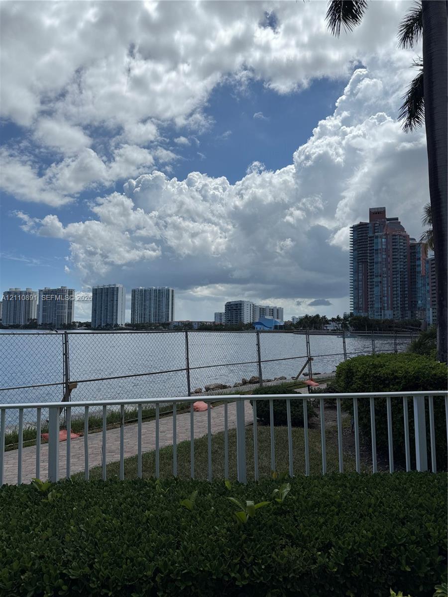 Waterfront grounds featuring a paved walkway, manicured hedging, metal fencing, and perimeter palms