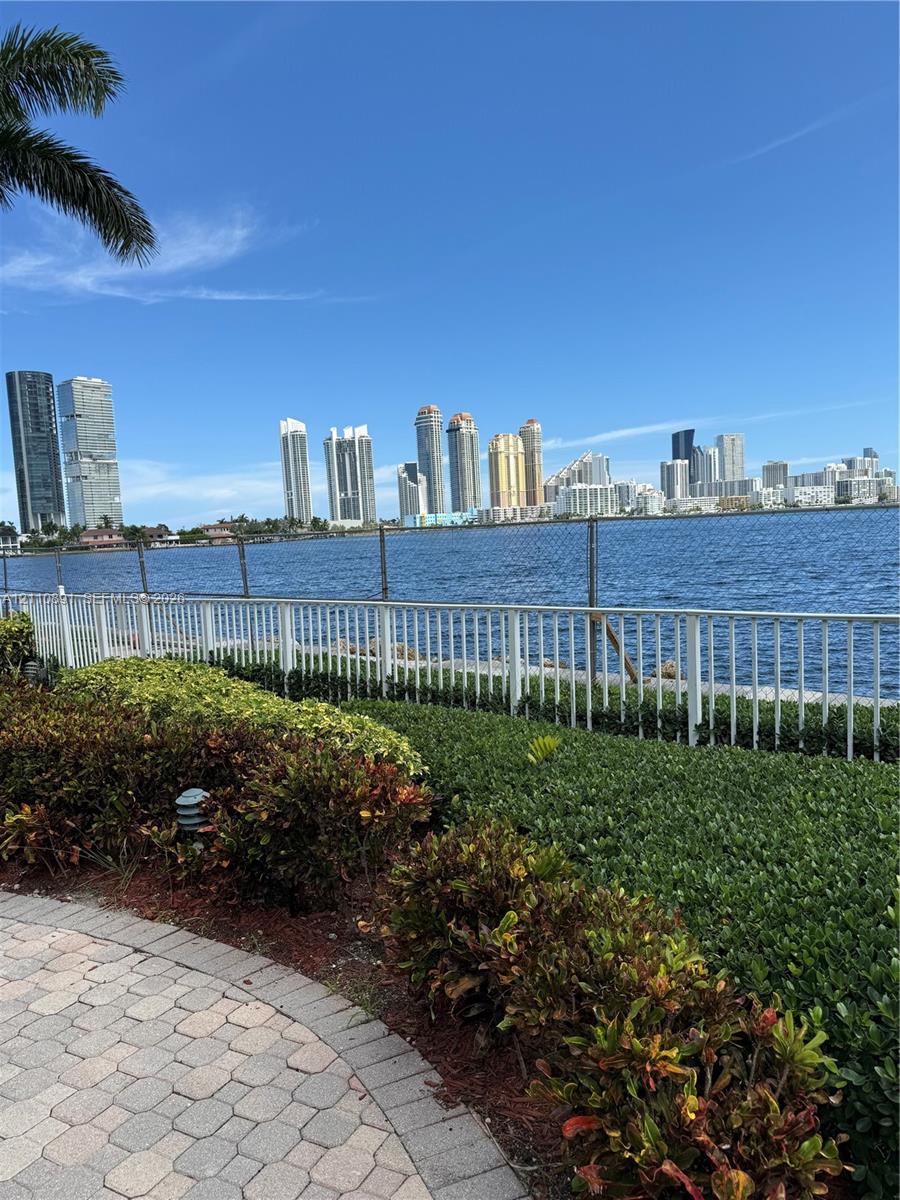 Waterfront property featuring a paved stone walkway, landscaped shrubbery, a white perimeter fence, and a skyline backdrop