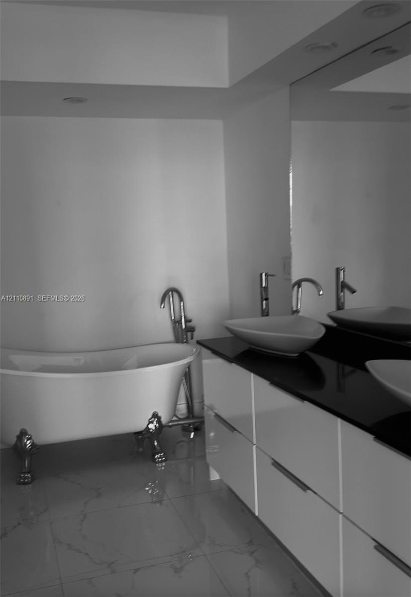 Freestanding clawfoot tub with floor-mounted faucet, dual vessel sinks, dark stone countertop, high-gloss white vanity cabinetry, and large-format tile flooring