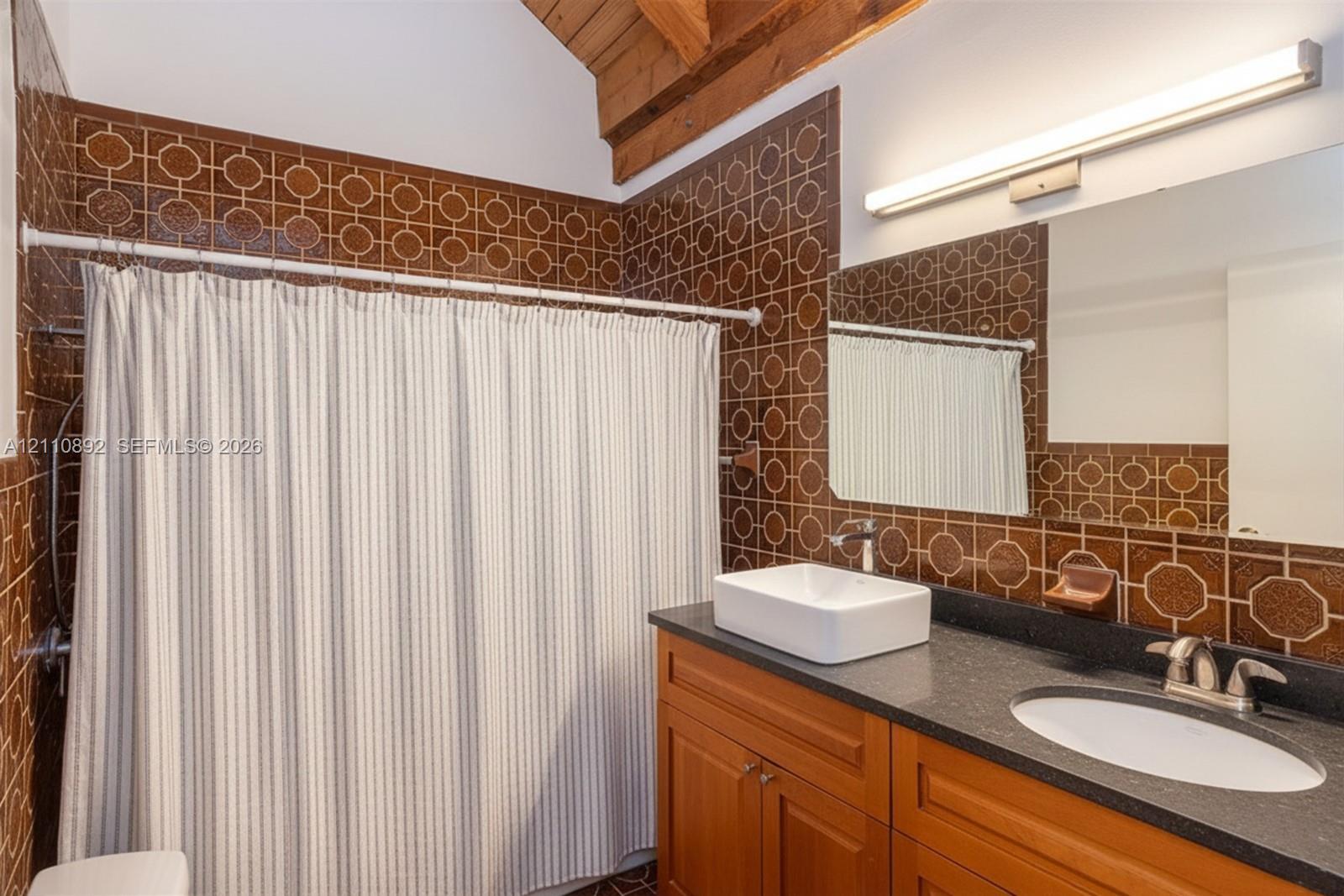 Bathroom featuring geometric-patterned wall tiling, a wood-finish vanity with a dark countertop, a rectangular vessel sink, and an exposed wood beam ceiling