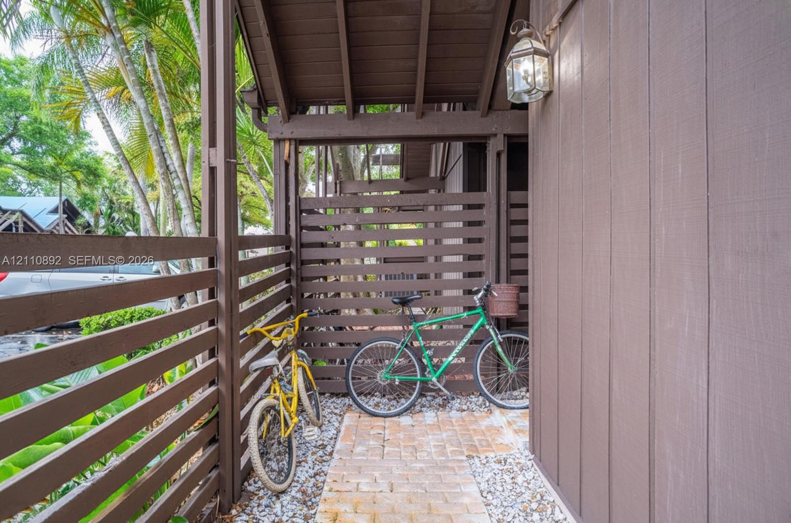 Covered entryway featuring vertical wood siding, horizontal slat privacy fencing, brick paver ground surface, and an exterior wall-mounted carriage-style light fixture
