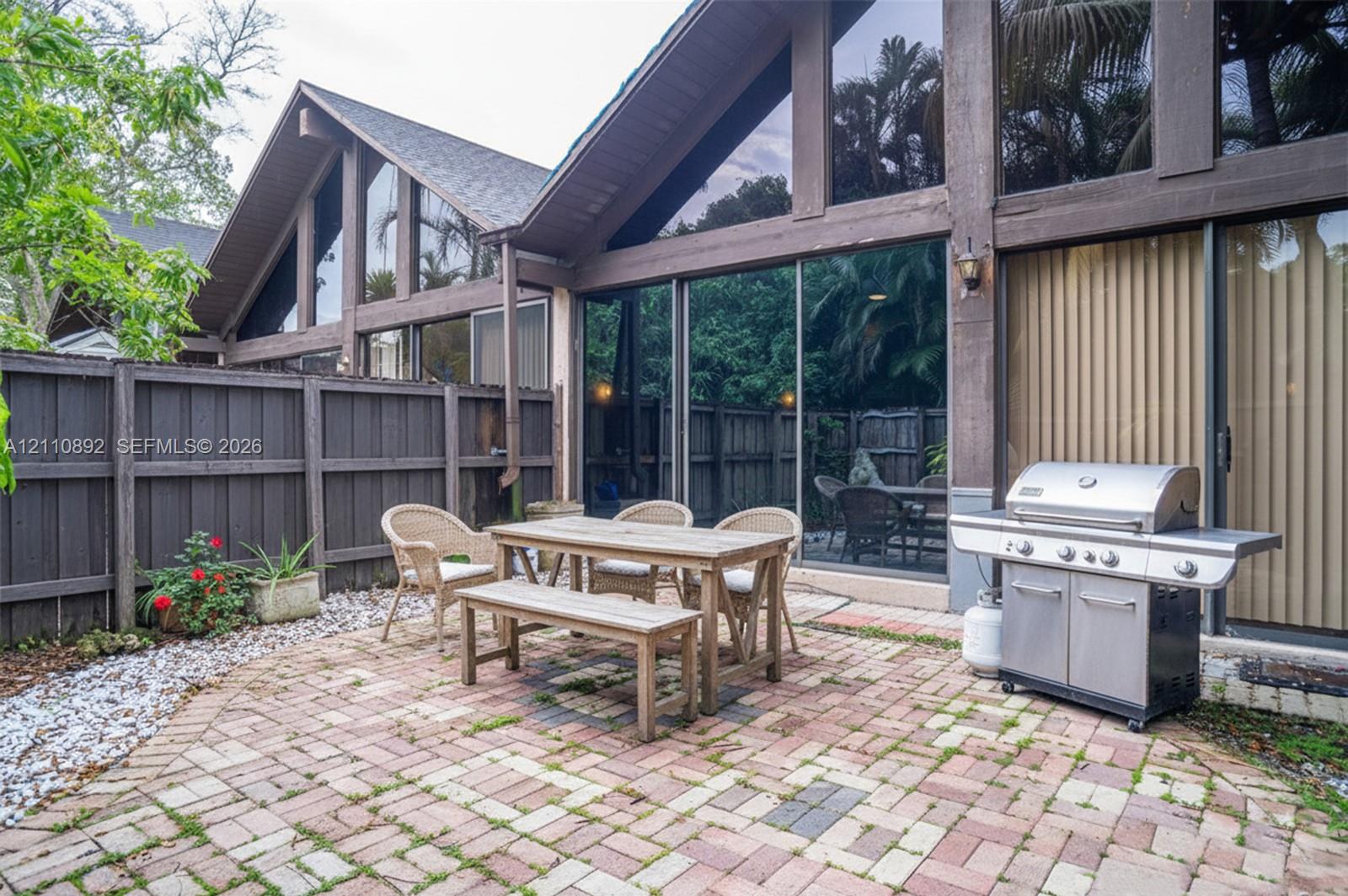 Paved brick patio featuring floor-to-ceiling glass paneling, A-frame architectural design, exterior wood-beam framing, and a perimeter wood fence Paved brick patio featuring floor-to-ceiling glass paneling, A-frame architectural design, exterior wood-beam framing, and a perimeter wood fence
