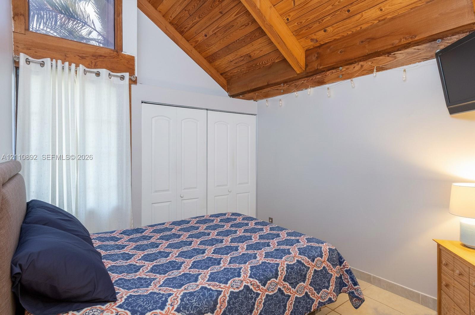 Vaulted ceiling with exposed wood beams, natural wood window trim, tile flooring, and built-in closet with double doors