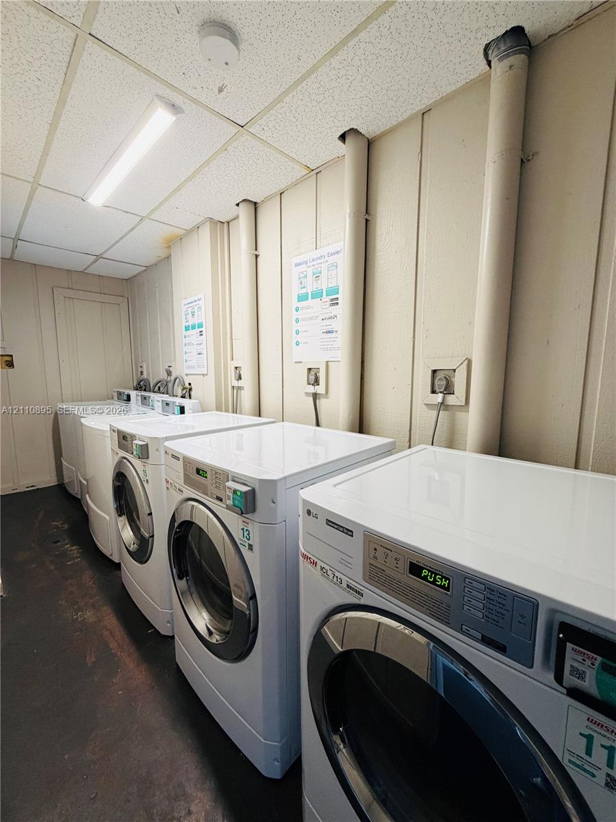 Laundry facility featuring multiple front-loading washers and dryers, vertical paneling, and drop-ceiling lighting