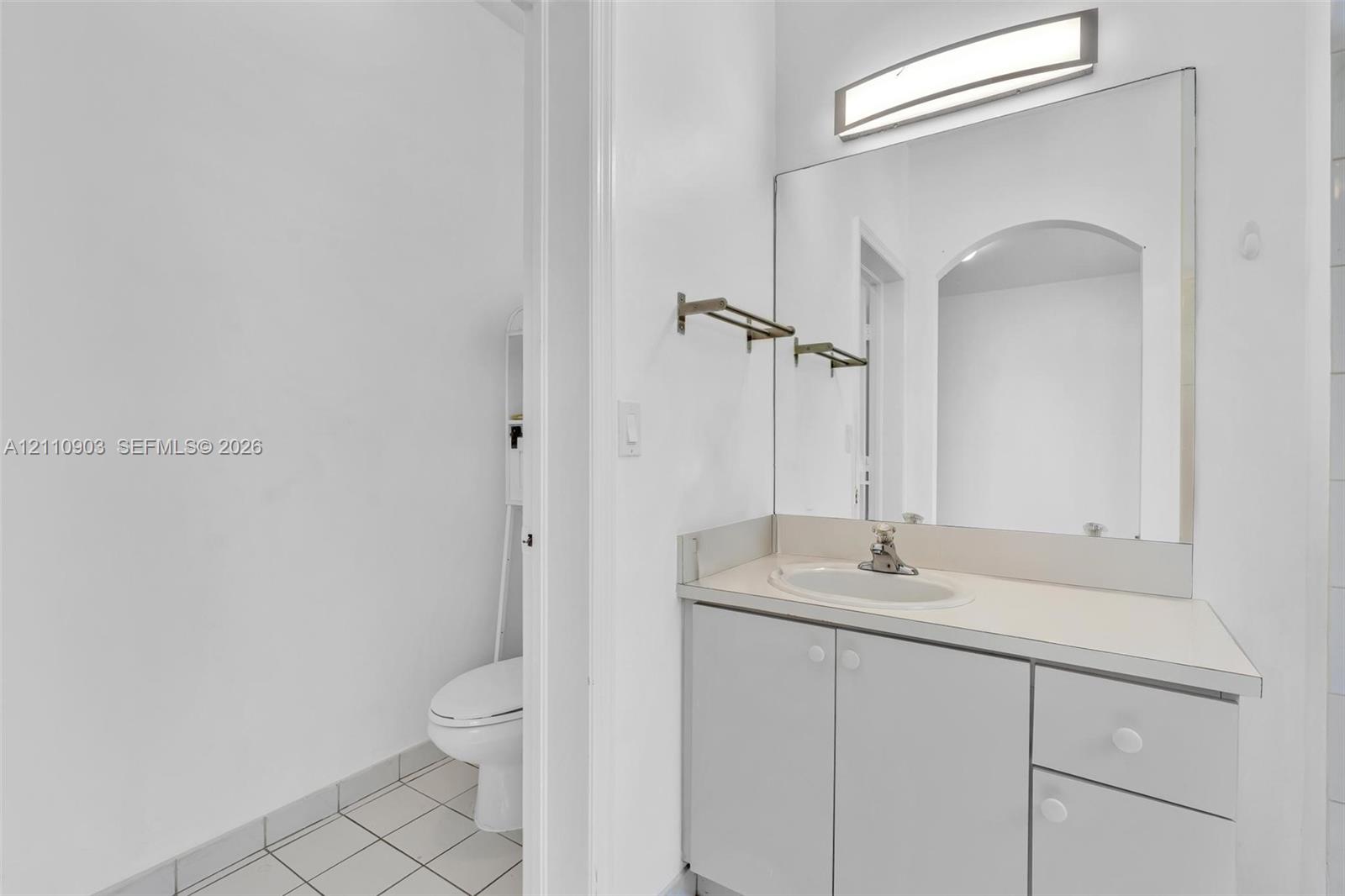 Bathroom vanity featuring light-toned cabinetry, an integrated sink, a full-width mirror, and a wall-mounted light fixture, paired with tile flooring and a standard-height toilet