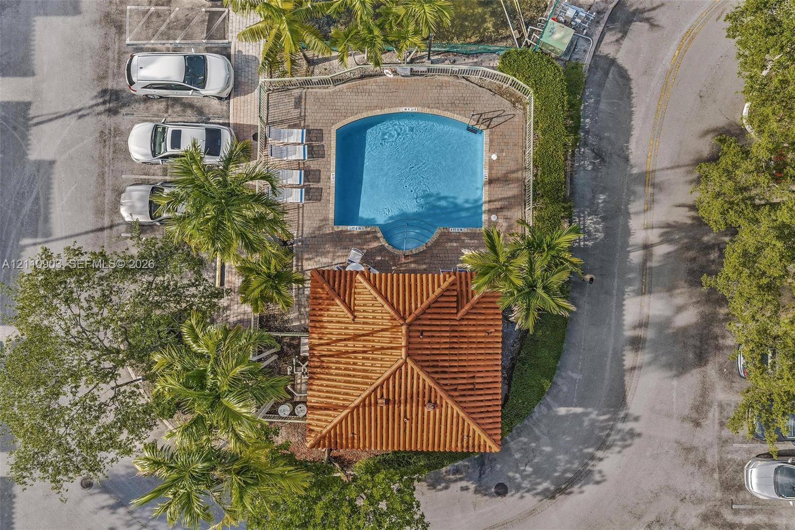 Aerial perspective of a community pool area featuring a curved basin, paver patio, terracotta-style roof structure, and mature palm trees