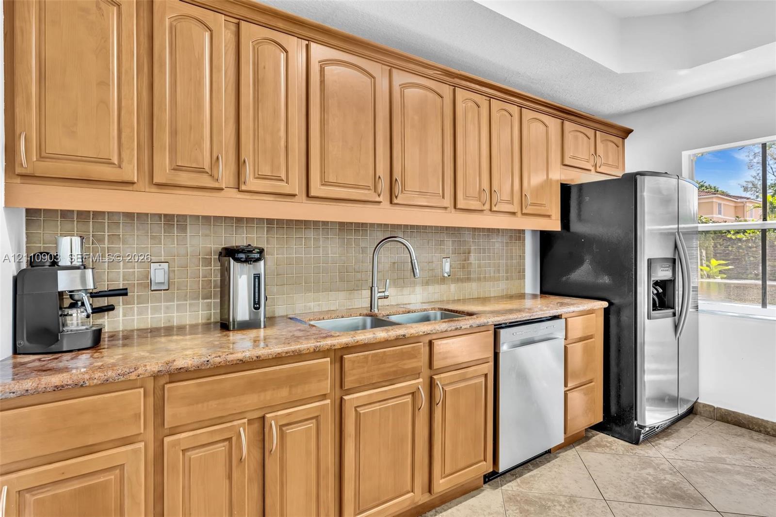 Kitchen featuring light-tone wood cabinetry, stone-surface countertops, mosaic tile backsplash, stainless steel dishwasher, and tile flooring