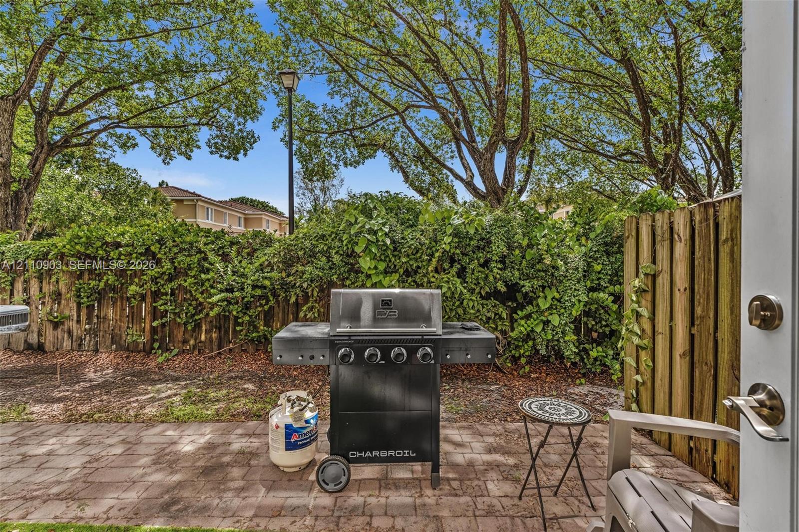 Paved patio area enclosed by a wood privacy fence, featuring mature shade trees and exterior lighting