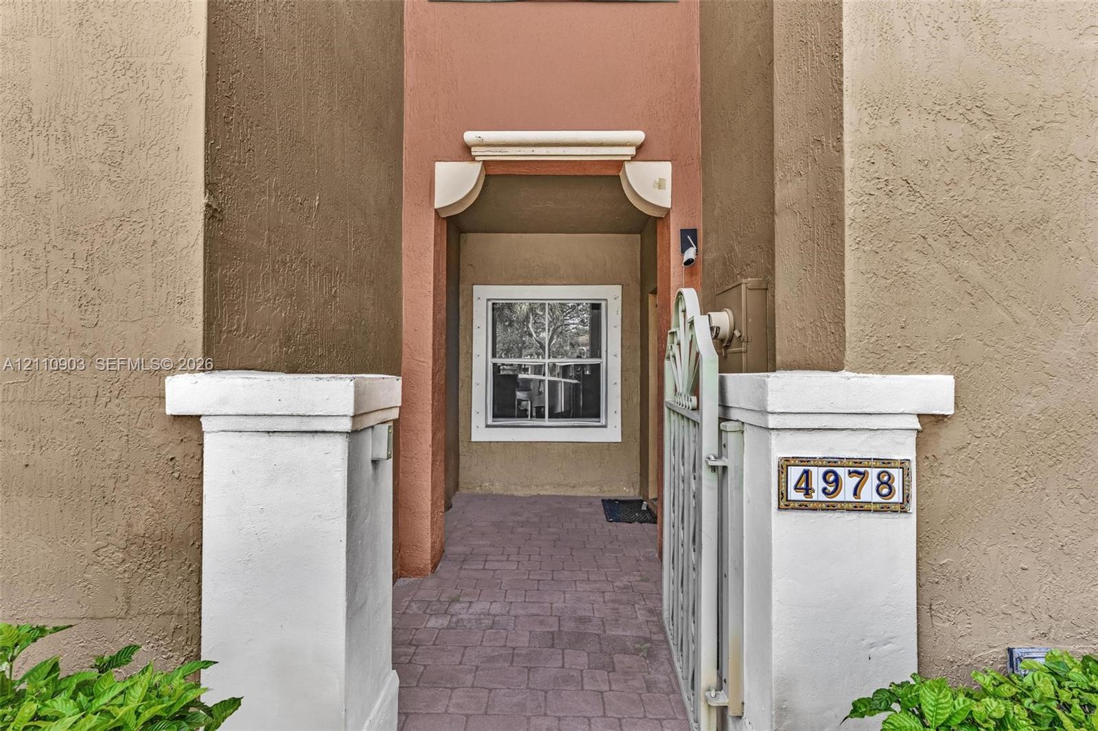 Stucco exterior entrance featuring white pillar bases, decorative metal gate, paver flooring, and arched architectural detailing