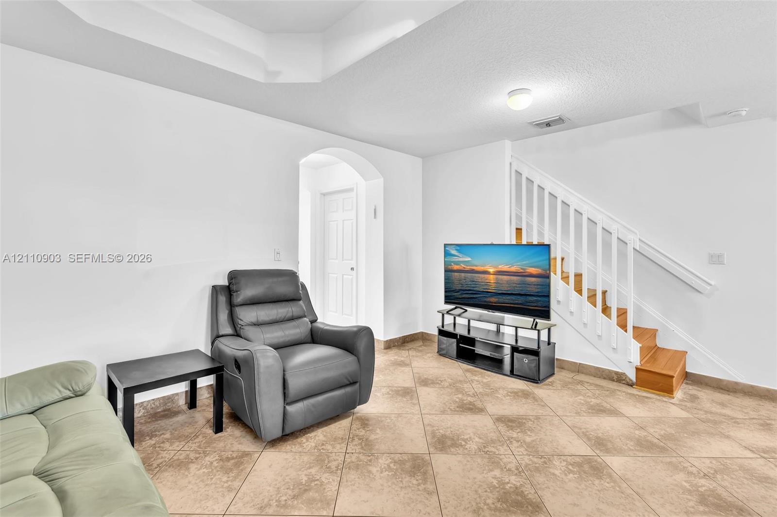 Interior space featuring a tray ceiling, large-format tile flooring, a white staircase with a wooden handrail, and an arched entryway