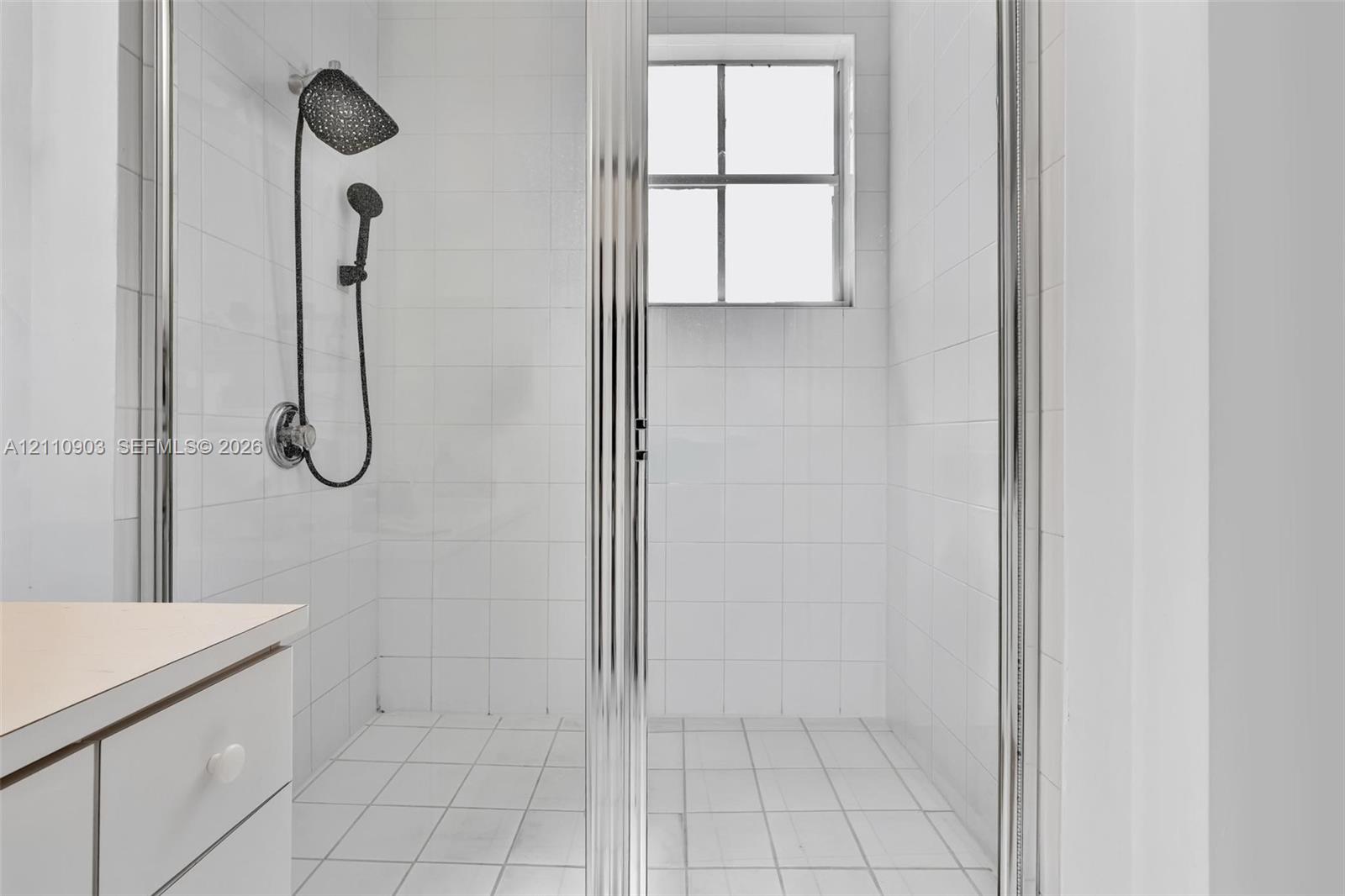 Walk-in shower enclosure with white tiled walls, square-grid tile flooring, chrome-framed glass door, and a dual-head shower fixture