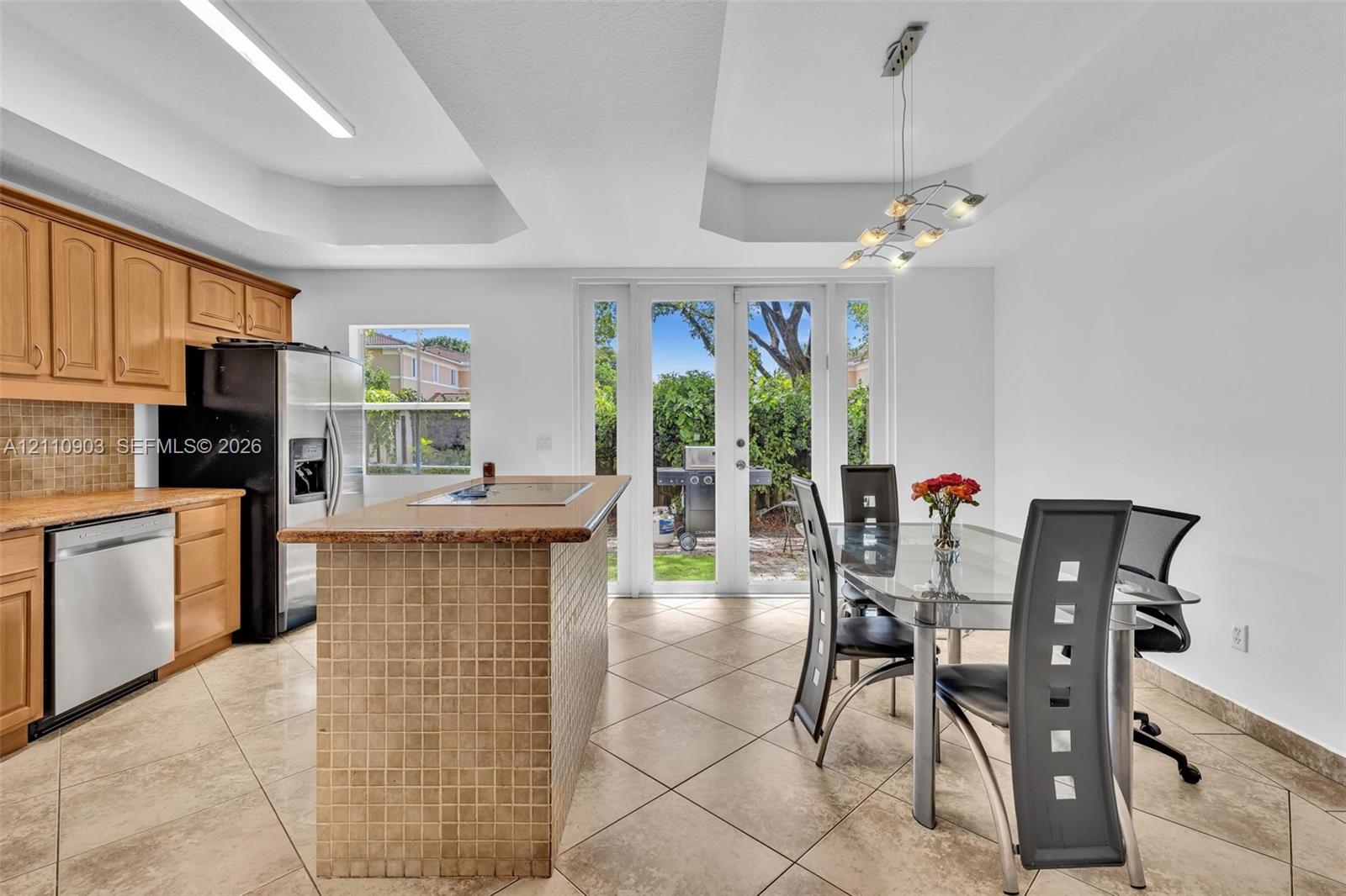 Kitchen featuring wood cabinetry, tiled island with a cooktop, stainless steel dishwasher, tray ceiling, and double glass doors