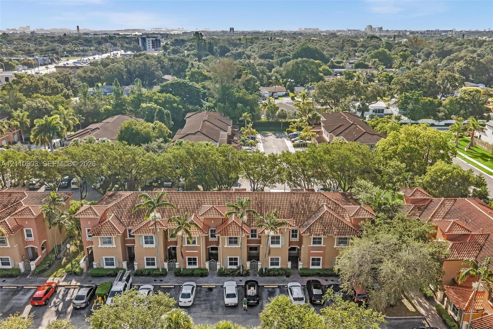 Aerial perspective of a multi-unit residential complex featuring a terracotta-style tile roof, stucco exterior, assigned parking, and mature surrounding tree canopy