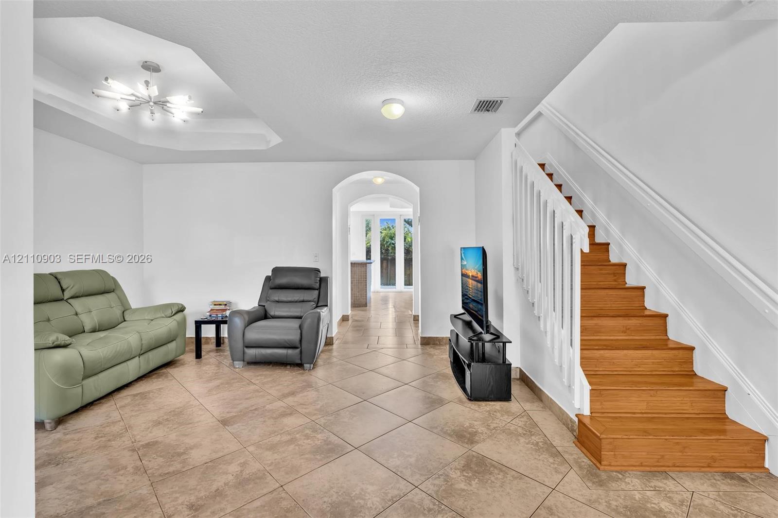 Tiled flooring, tiered tray ceiling with modern chandelier, staircase with white balusters and wood treads, and arched doorway
