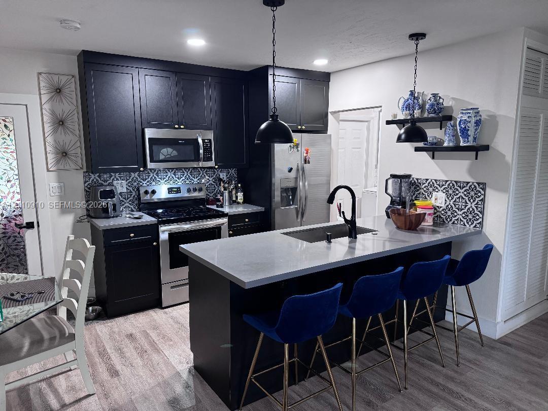 Kitchen featuring charcoal-toned shaker cabinetry, quartz countertops, patterned tile backsplash, stainless steel appliances, and a central island with an integrated sink and matte black faucet