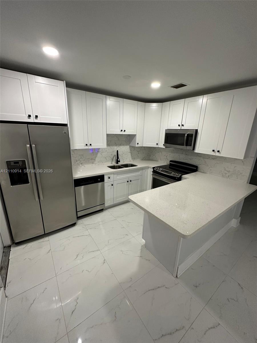 Kitchen featuring white shaker-style cabinetry, stone-finish countertops, stainless steel appliances, a peninsula, and large-format tile flooring with veining