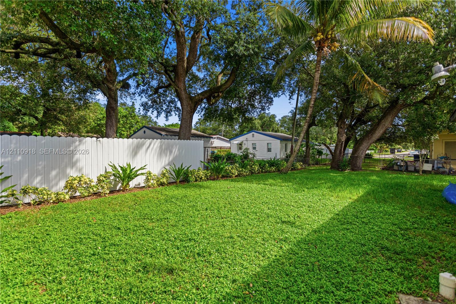 Spacious backyard featuring a white vinyl privacy fence, mature trees, established palms, and a manicured lawn