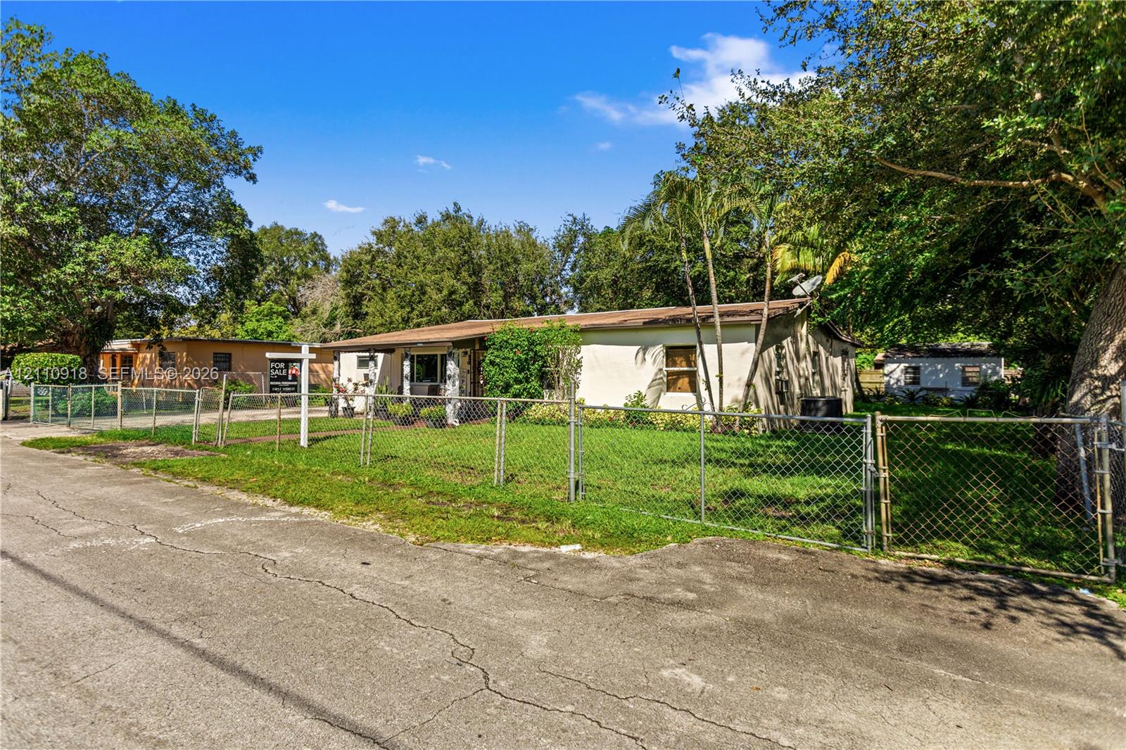 Single-story exterior with a stucco finish, metal roof, chain-link perimeter fencing, and mature trees