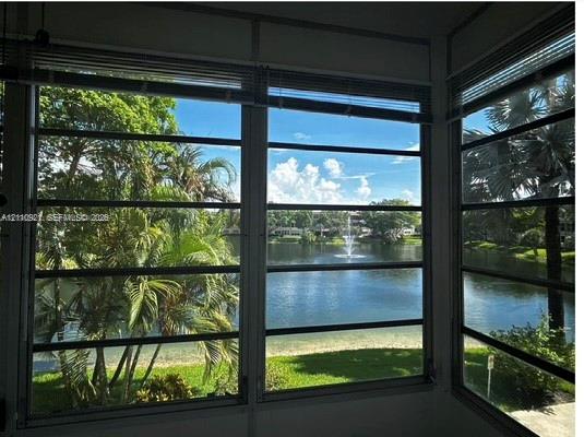 Enclosed space featuring aluminum-framed louvered windows, horizontal blinds, and a direct perspective of a pond with a central water fountain, mature palm trees, and landscaped grounds Enclosed space featuring aluminum-framed louvered windows, horizontal blinds, and a direct perspective of a pond with a central water fountain, mature palm trees, and landscaped grounds