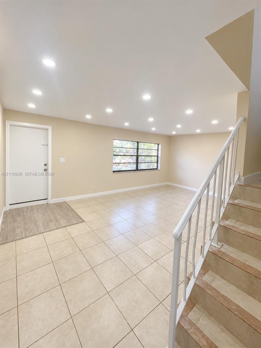 Open-concept interior featuring light-toned tile flooring, recessed ceiling lighting, a multi-pane window, and a staircase with a white metal railing
