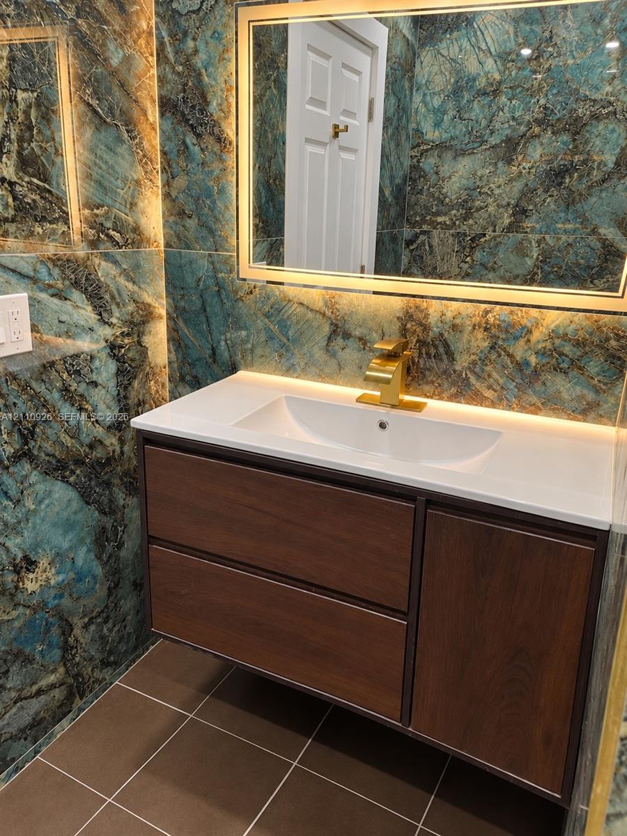 Wall-mounted wood-finish vanity with a rectangular basin, stone-composite countertop, metallic gold-tone faucet, and an integrated LED-backlit mirror framed by polished stone tile walls