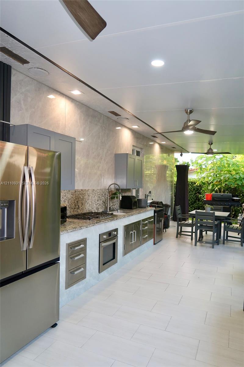 Outdoor kitchen featuring stone countertops, built-in stainless steel appliances, cabinetry with light gray finish, recessed ceiling lighting, and large-format tile flooring