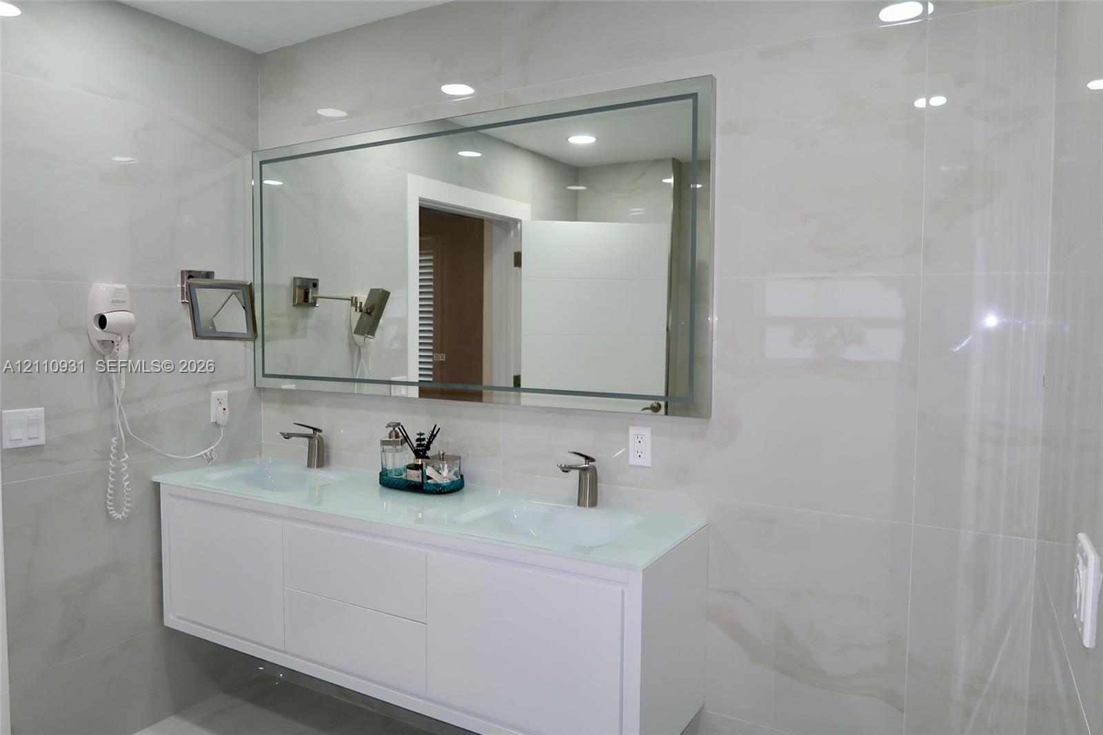 Wall-mounted white dual-sink vanity featuring a glass countertop, large rectangular mirror with integrated lighting, wall-mounted adjustable magnification mirror, and large-format neutral-toned tile wall surfacing