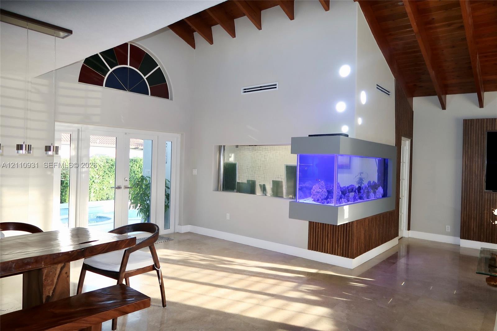 Vaulted ceiling featuring exposed wood beams, built-in recessed aquarium, arched accent window, glass-paneled double doors, and polished stone flooring