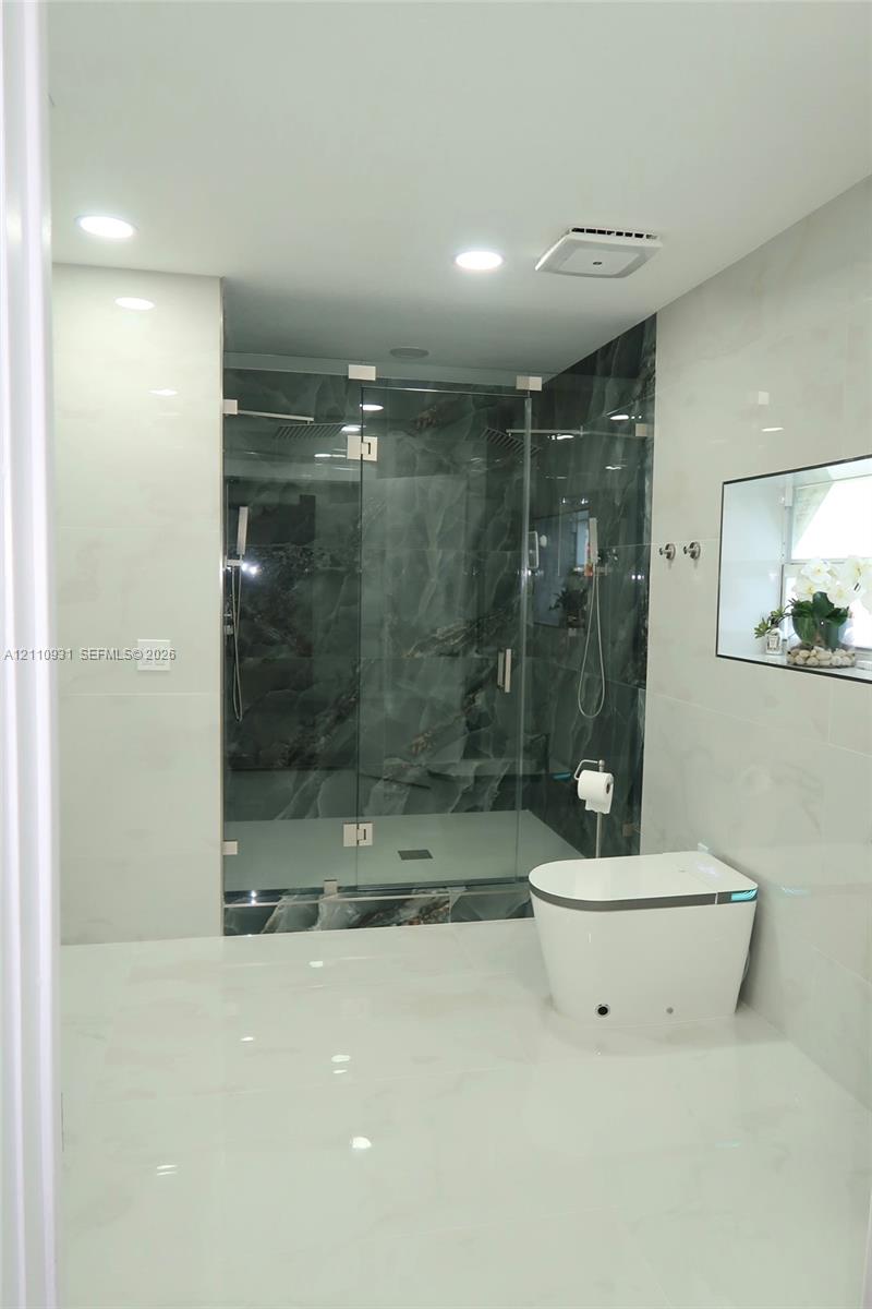 Walk-in glass-enclosed shower with dark stone-patterned tile, white floor-to-ceiling wall tile, recessed lighting, and a modern low-profile toilet