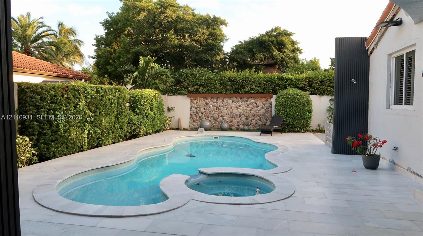 Freeform swimming pool and integrated spa, light-toned stone decking, and decorative stone masonry feature wall surrounded by privacy hedging