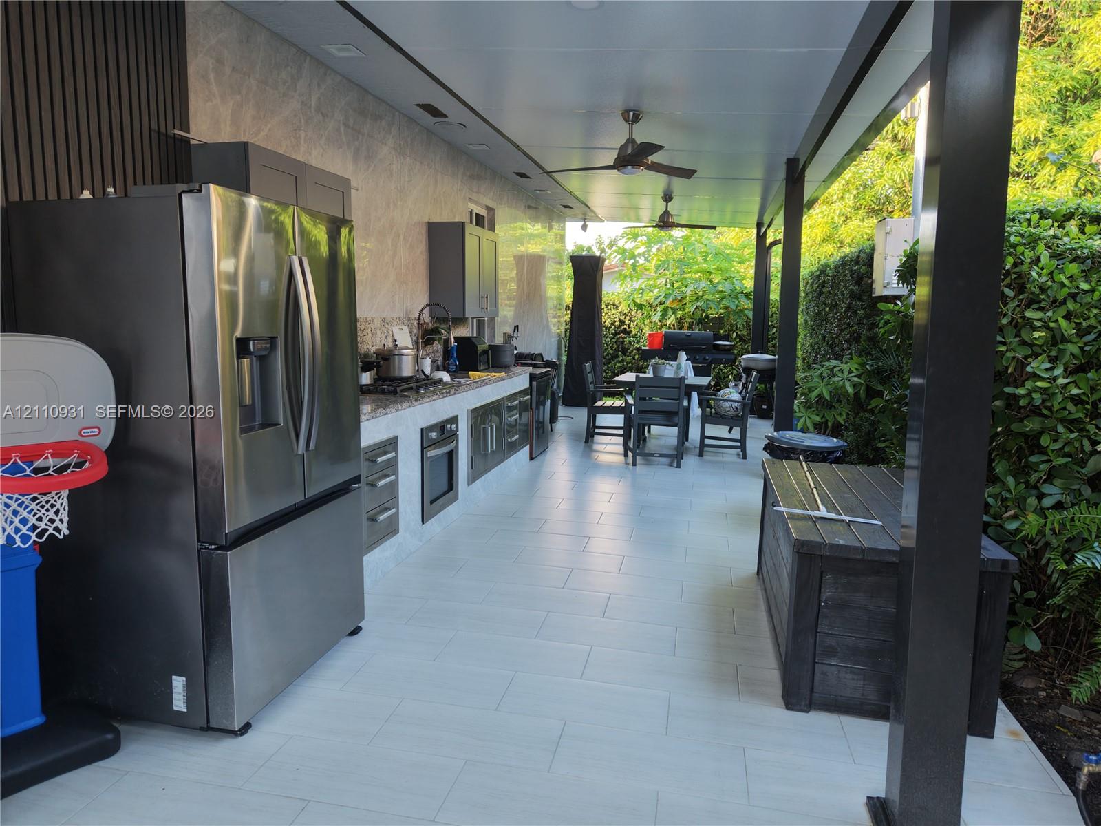 Covered outdoor kitchen featuring tile flooring, stainless steel appliances, stone-finish backsplash, built-in cabinetry, and ceiling fans