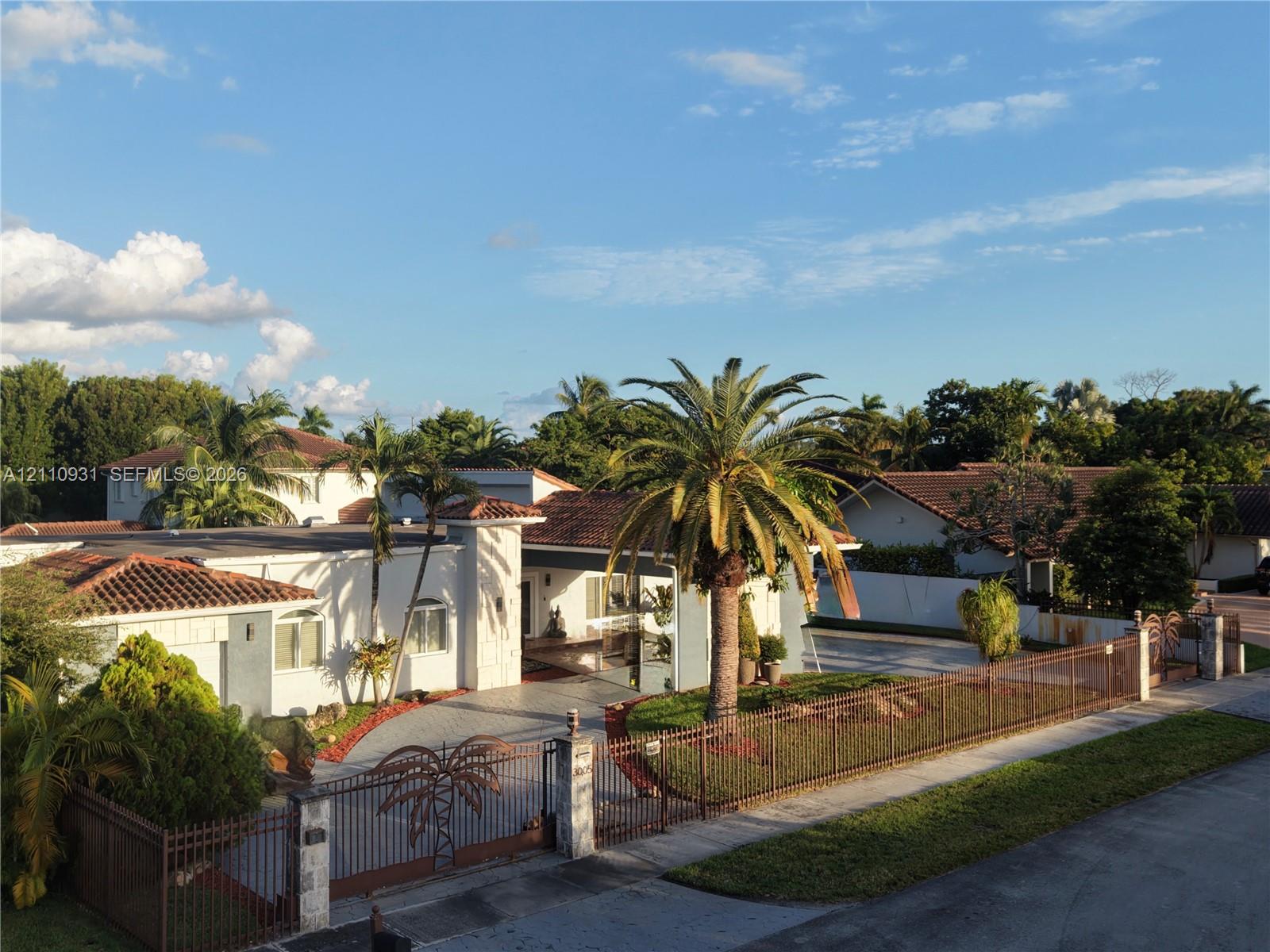 Single-story residence featuring a terracotta tile roof, decorative metal perimeter fencing, paved driveway, and mature palm tree landscaping