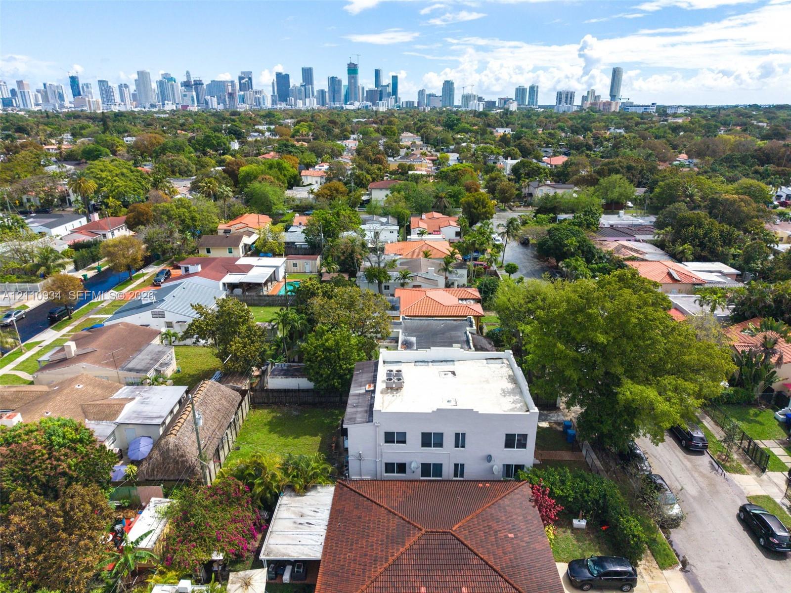 Aerial perspective of a multi-story building featuring a flat roof and surrounding mature landscaping, positioned within a suburban environment with a prominent city skyline backdrop