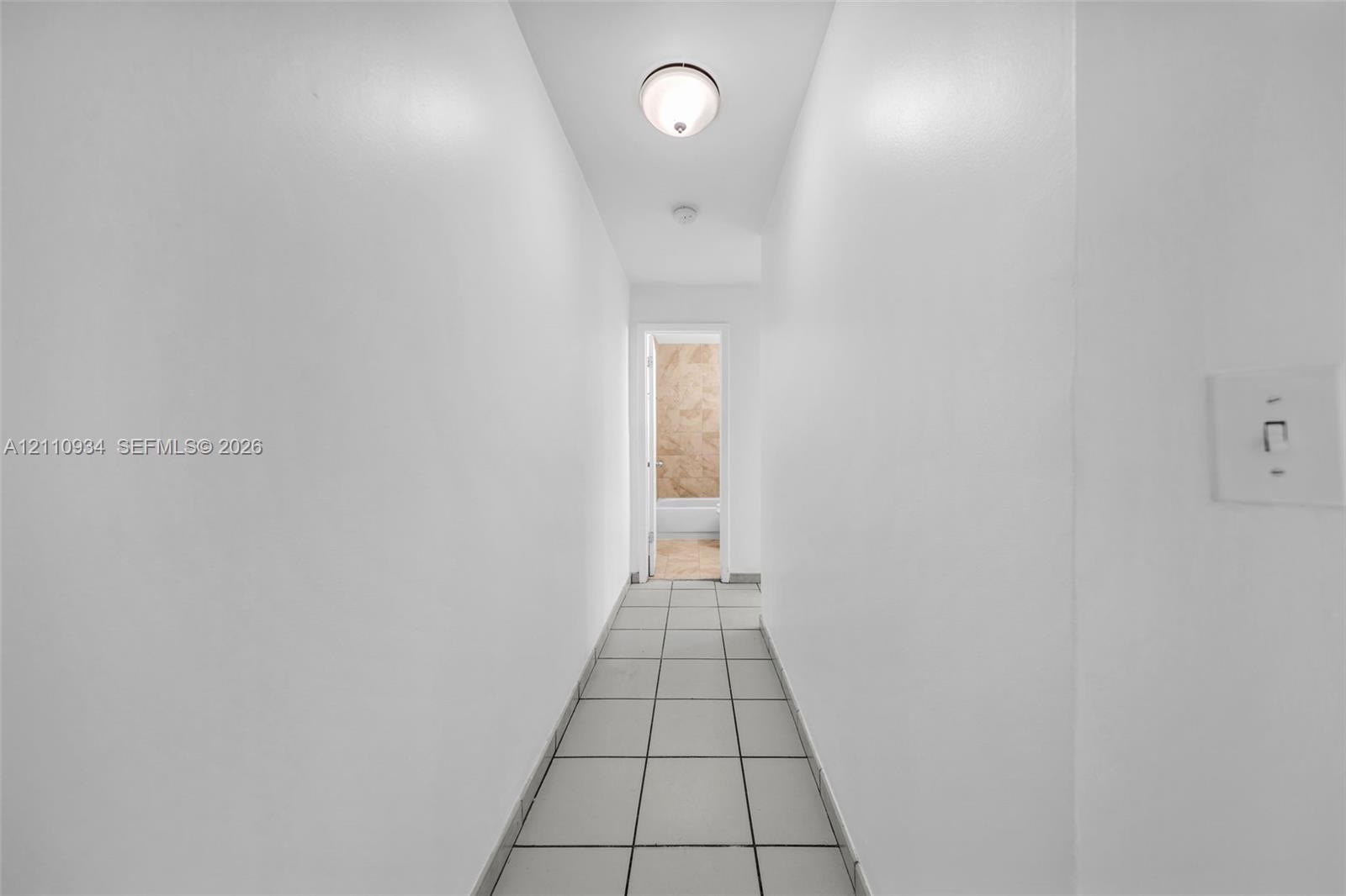 Hallway featuring neutral-toned tile flooring, smooth wall surfaces, and a flush-mount overhead light fixture