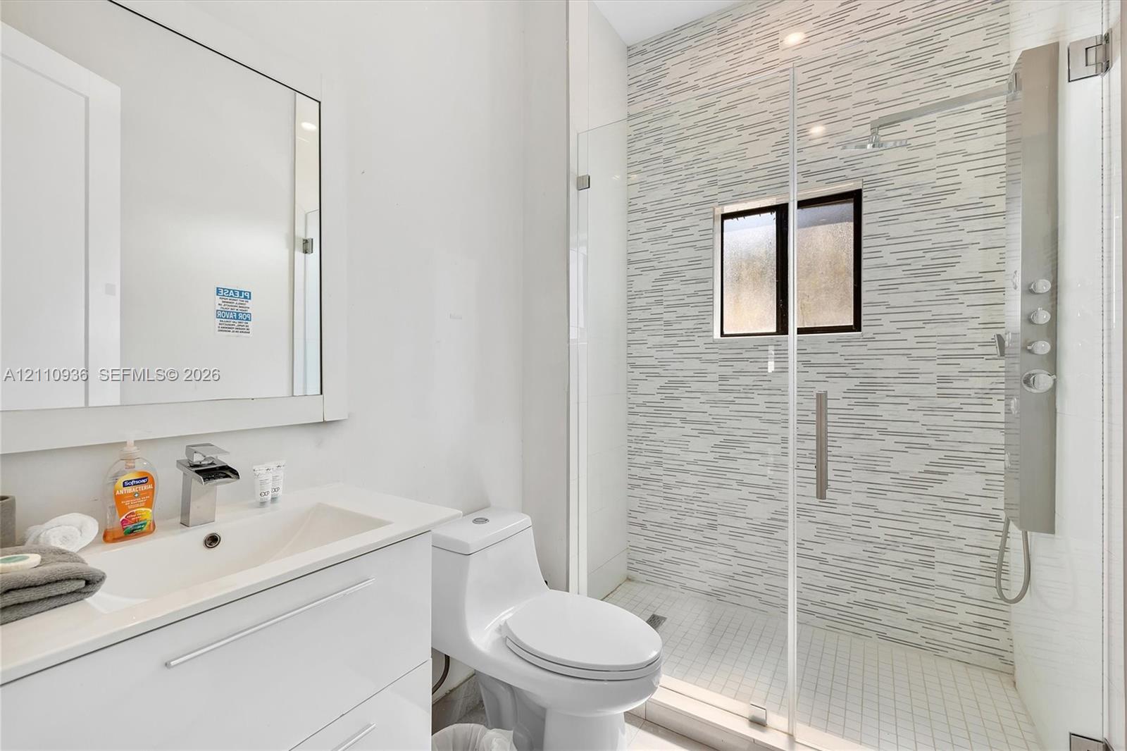 Modern bathroom featuring a white vanity with an integrated countertop, wall-mounted mirror, walk-in shower with a glass enclosure, patterned tile shower wall, and an integrated shower panel system Modern bathroom featuring a white vanity with an integrated countertop, wall-mounted mirror, walk-in shower with a glass enclosure, patterned tile shower wall, and an integrated shower panel system
