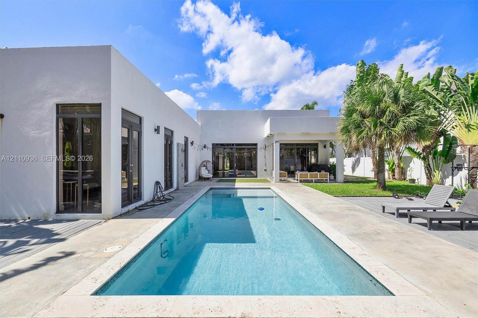 Modern exterior with smooth stucco finish, rectangular swimming pool with stone coping, concrete patio, covered outdoor area, and established palm trees