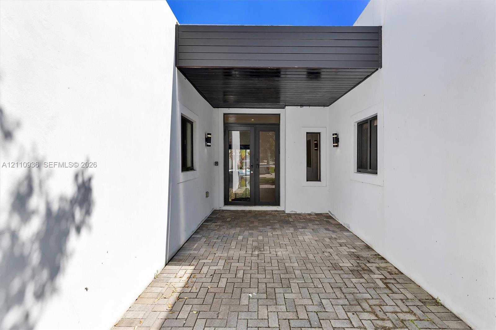 Covered entryway featuring a herringbone-patterned paver floor, minimalist smooth-stucco exterior walls, dark wood-finish ceiling paneling, glass-paneled double doors, and wall-mounted exterior sconces Covered entryway featuring a herringbone-patterned paver floor, minimalist smooth-stucco exterior walls, dark wood-finish ceiling paneling, glass-paneled double doors, and wall-mounted exterior sconces