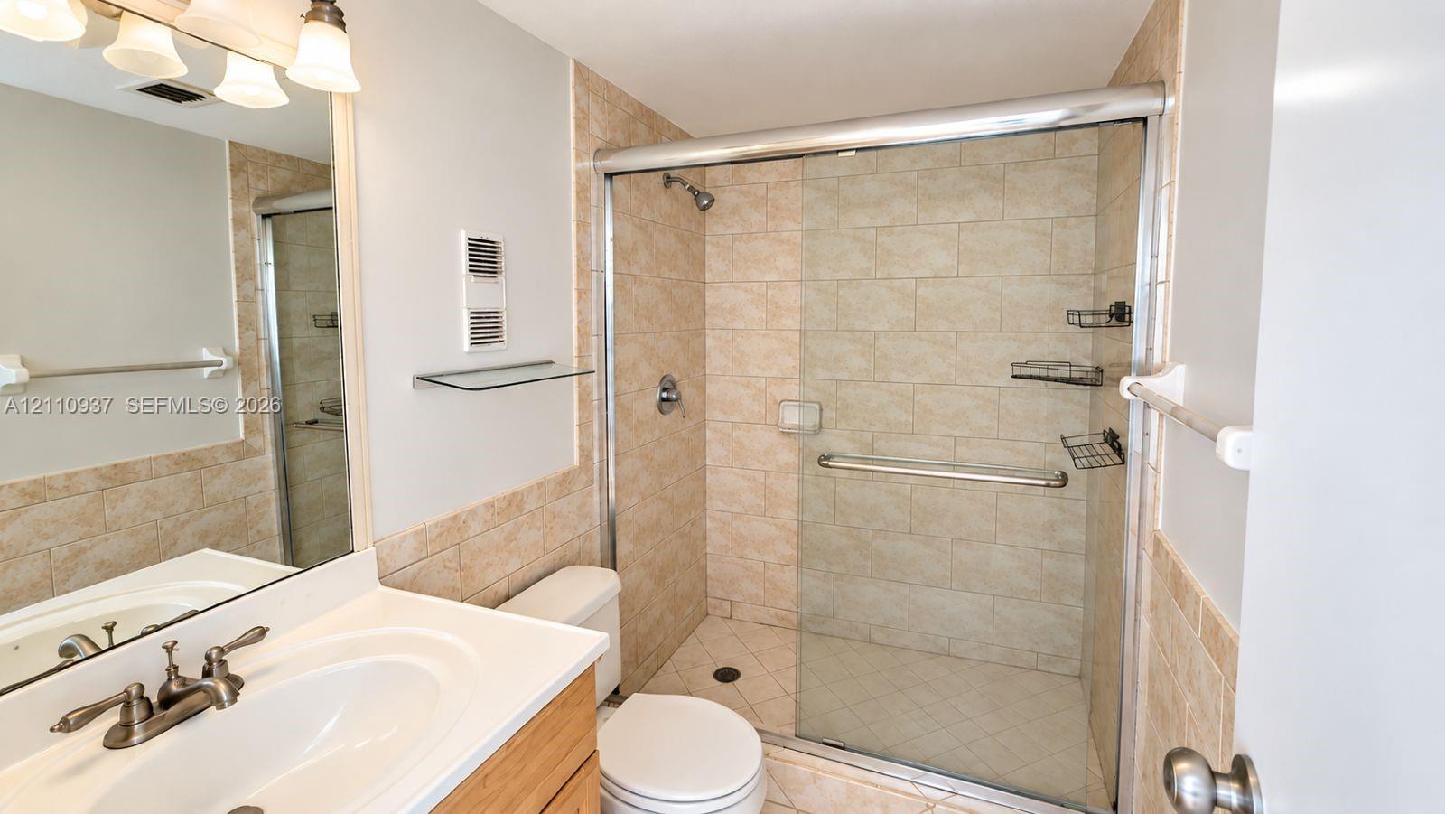 Bathroom featuring a vanity with integrated sink, stone-finish tile shower surround, sliding glass shower door, and wall-mounted vanity lighting
