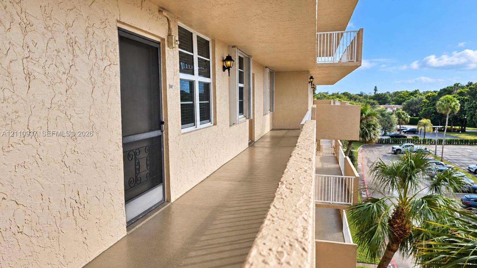 Exterior walkway featuring a stucco facade, metal screen door, white-framed multi-pane windows, wall-mounted coach lighting, and a white railing