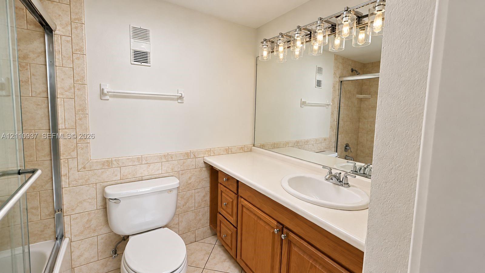 Bathroom featuring a wood vanity with a solid-surface countertop, an integrated oval sink, ceramic tile wainscoting, and a multi-light fixture over a wall-to-wall mirror