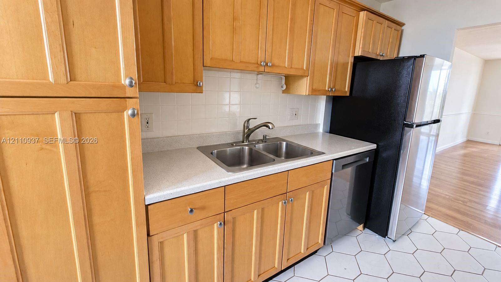 Kitchen featuring light wood cabinetry, white tile backsplash, solid-surface countertops, dual-basin stainless steel sink, and stainless steel dishwasher