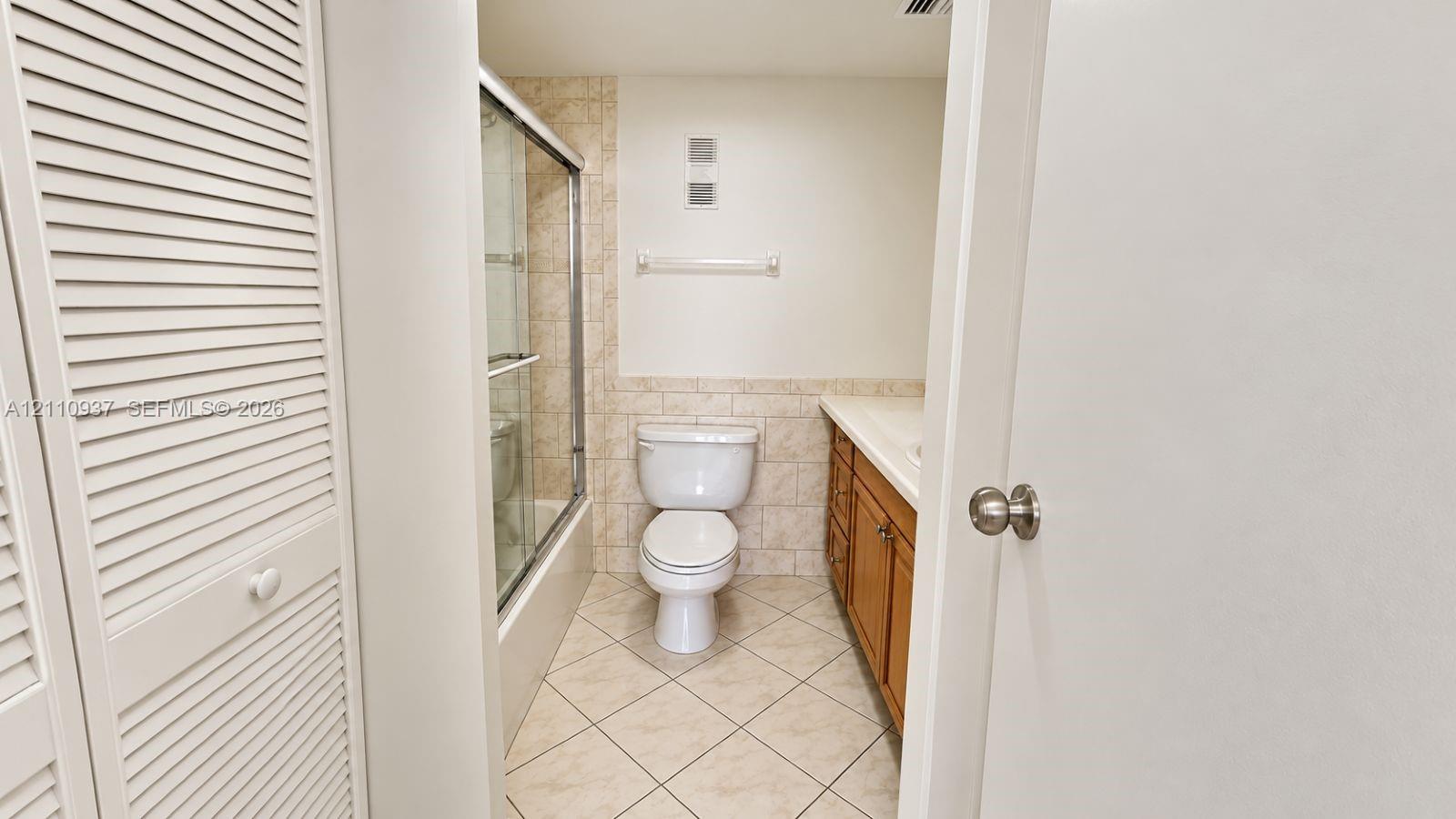 Bathroom featuring wood-finish cabinetry, neutral-toned tiled flooring laid on a diagonal, a glass-enclosed shower with tiled walls, and louvered closet doors